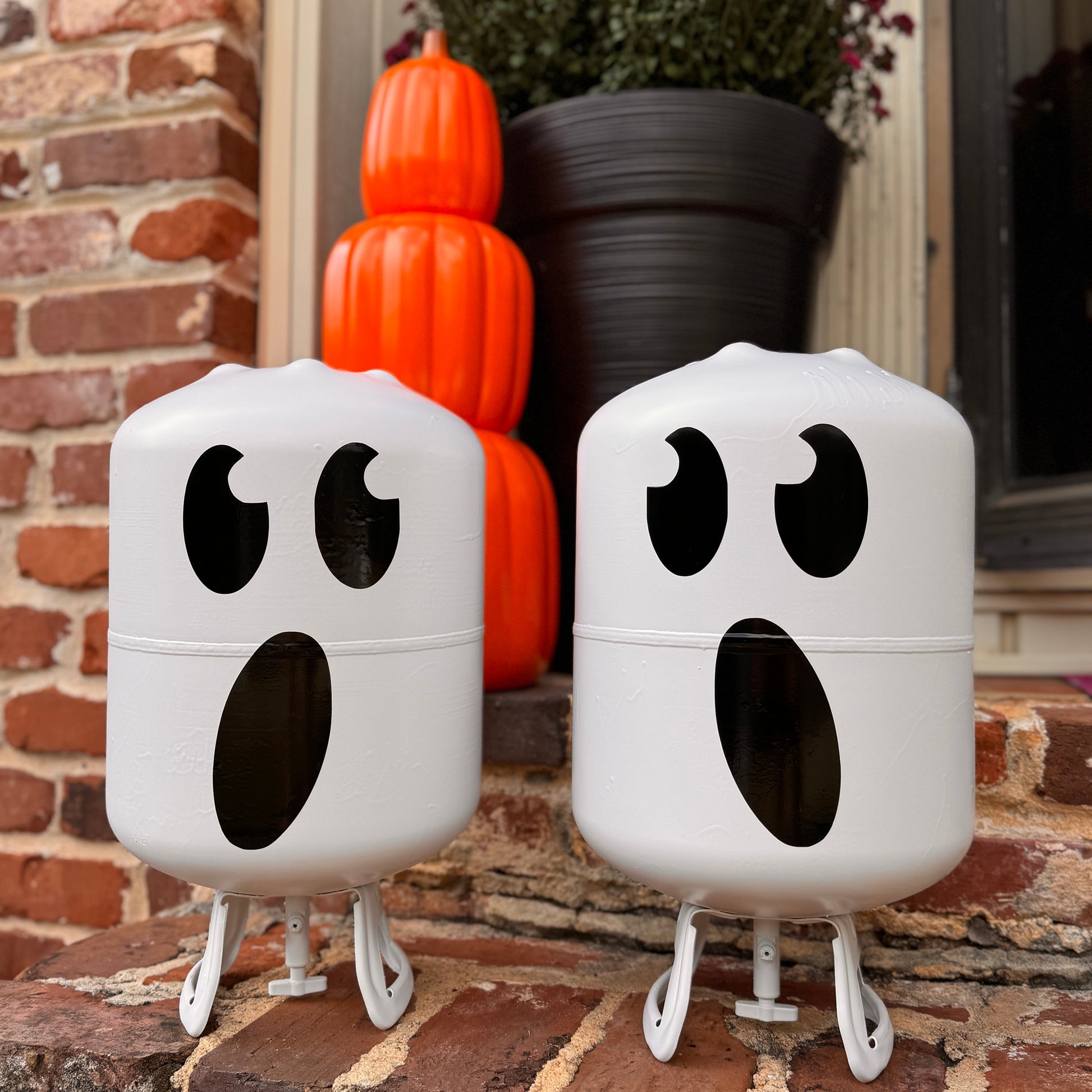 Two ghost-shaped outdoor lights on a brick surface with pumpkins and flowers in the background.